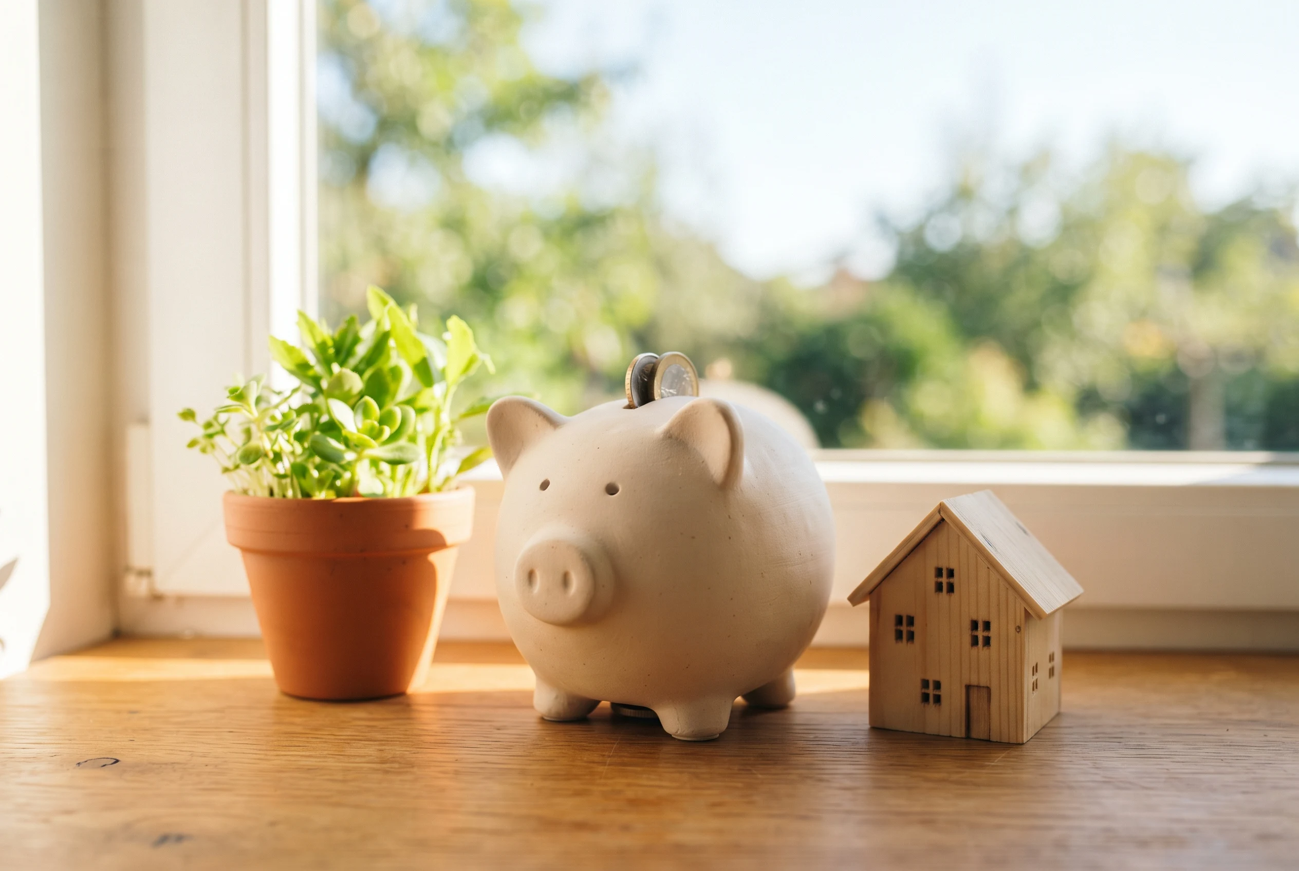 Sparschwein mit grünen Blättern neben einem Holzhaus-Modell in sonnigem Licht, Symbol für die Finanzierung der Wärmepumpe im Altbau